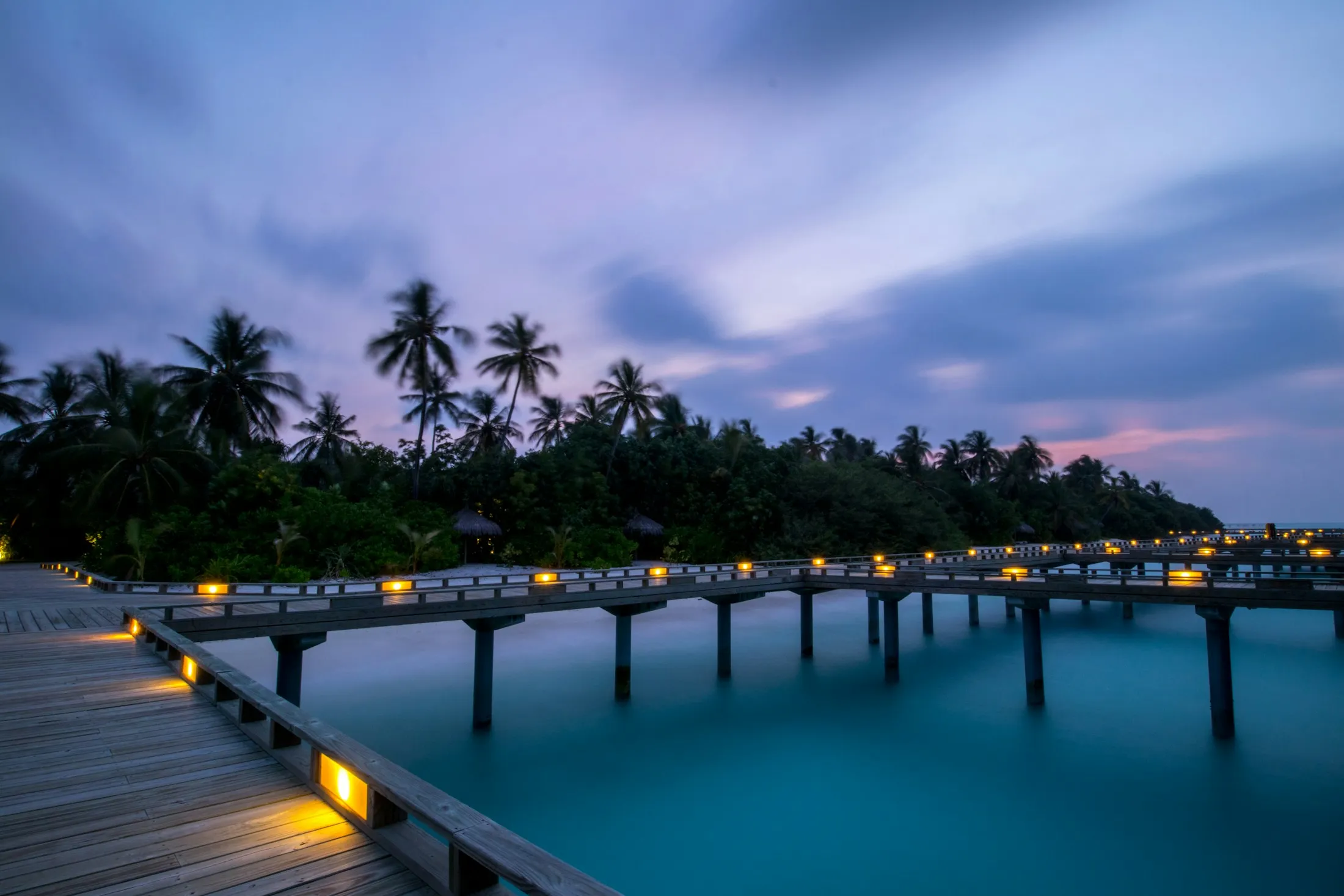 An illuminated wooden jetty leading to overwater villas at dusk